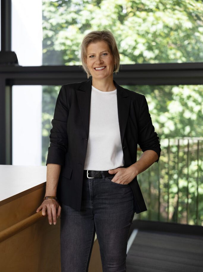 Petra Kamper, smiling, wearing a dark blazer in front of a large window with trees.