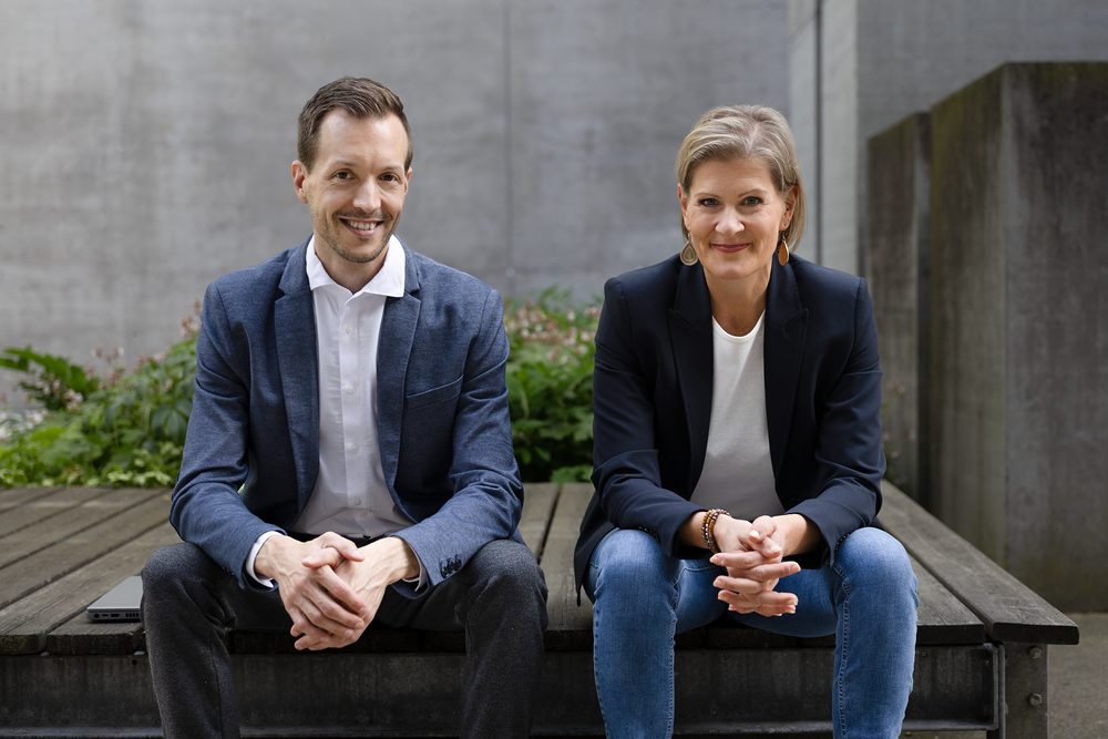 Stefan Kirchsteiger and Petra Kamper sitting side by side on a wooden bench in front of a concrete wall.