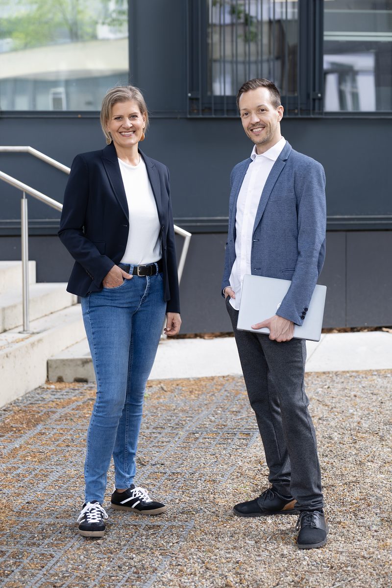 Petra Kamper and Stefan Kirchsteiger, holding a laptop, both standing in front of a building with a dark facade.
